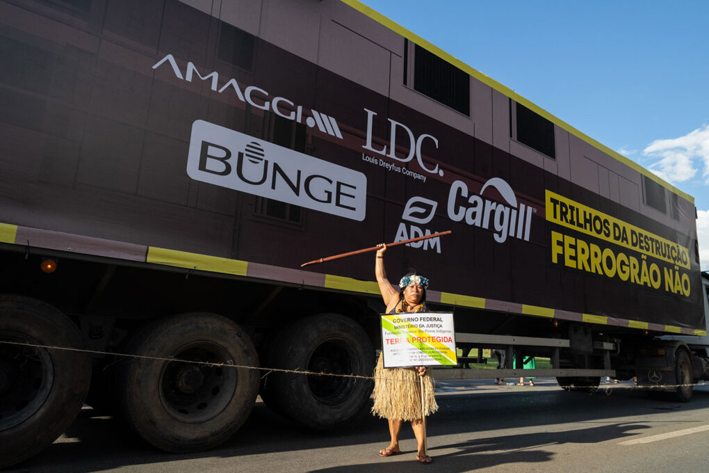 An activist from the #NoFerragão Allinace Stands in front of a train covered in corporate logos. Photo credit: Felipe Beltram.